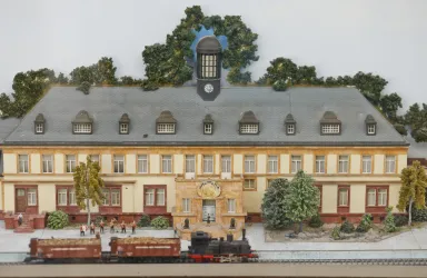 Ein detailliertes Modell eines Bahnhofs mit einem großen Uhrenturm. Vor dem Gebäude stehen Züge und Bäume, umgeben von einer kleinen Parklandschaft.