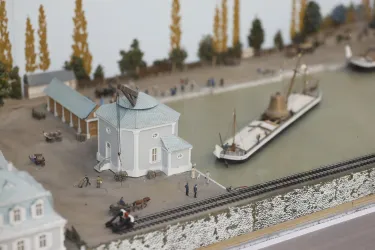 Eine detaillierte Modellansicht eines Hafenbereichs mit einem Schiff und einer kleinen Hafenanlage. Umgeben ist die Szene von Bäumen und Figuren, die den Alltag im Hafen darstellen.