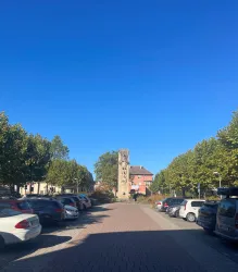 Eine Straße mit autostellenden Parkplätzen und einer hohen Säule in der Mitte. Der Himmel ist klar und blau, und grüne Bäume säumen die Straße.