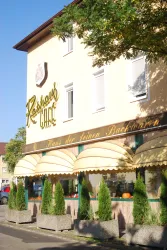 Ein helles Café mit der Aufschrift "Ressers Café". Vor dem Gebäude stehen große, grüne Pflanzen in Betonkübeln.