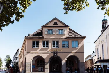 Ein historisches Gebäude mit einem schönen Dach und großen Fenstern. Vor dem Gebäude stehen Menschen und Bäume spenden Schatten.