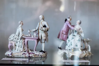 Zwei elegante Porzellanfiguren in einem Ausstellungsstück. Eine Figur zeigt eine Teeservice-Szene, die andere ein Paar im Gespräch.
