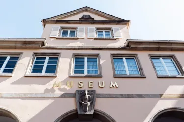 Das Bild zeigt die Fassade eines Museums mit mehreren Fenstern und einem stattlichen Schild, auf dem "MUSEUM" steht. Die Architektur ist schlicht und freundlich, unter einem klaren Himmel.