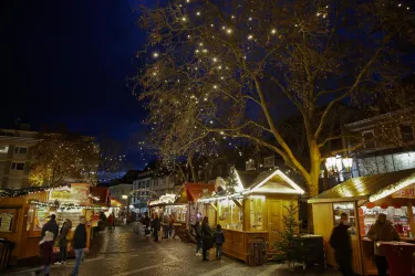 Ein festlicher Weihnachtsmarkt mit beleuchteten Ständen und einem großen Baum. Menschen schlendern bei Nacht durch die geschmückte Szenerie.