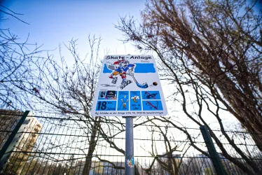 Ein Schild weist auf eine Skate-Anlage hin. Im Hintergrund sind kahle Bäume und ein blauer Himmel zu sehen.