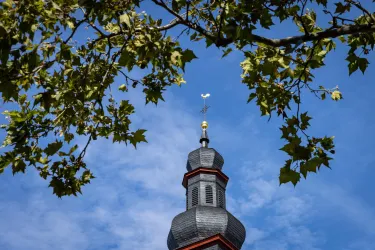 Ein Kirchturm ragt unter einem blauen Himmel empor, umgeben von grünen Blättern. Die Architektur ist traditionell und auffällig.
