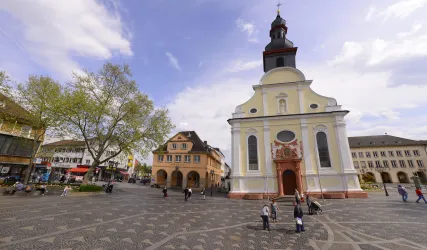 Eine beeindruckende Kirche steht auf einem Platz mit modernen und historischen Gebäuden. Die Bäume und der Himmel verleihen der Szene eine einladende Atmosphäre.