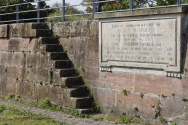 Eine steinerne Treppe führt zu einer Mauer mit einer Tafel. Die Tafel zeigt eingelassene Schriftzüge und ist von Gras umgeben.