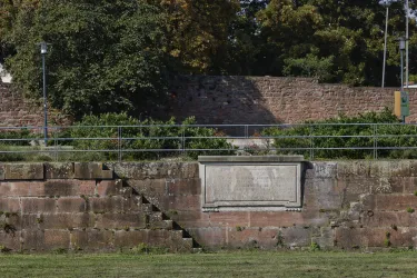 Ein historischer Mauerabschnitt mit einer hervorstehenden Tafel. Im Hintergrund sind Bäume und eine gewellte Mauer sichtbar.