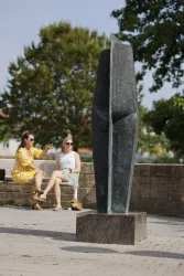 Eine moderne Skulptur steht im Vordergrund. Zwei Frauen sitzen dahinter und genießen die Zeit im Freien.