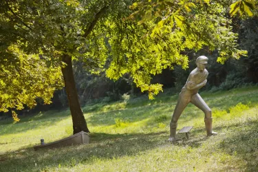 Eine silberne Skulptur eines Mannes steht in einem grünen Park. Im Hintergrund sind Bäume und Wiese zu sehen.