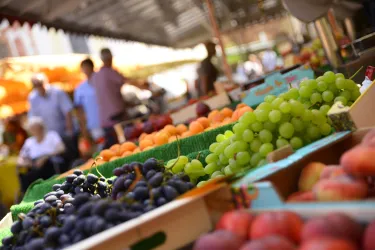 Ein bunter Markt mit verschiedenen Obstsorten wie Trauben, Äpfeln und Orangen. Im Hintergrund sind Menschen zu sehen, die den Markt besuchen.