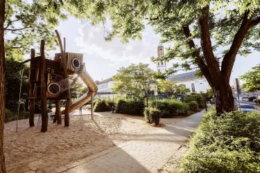 Ein Spielplatz mit einer Rutsche und Spielgeräten aus Holz. Umgeben von Bäumen und Grünflächen.