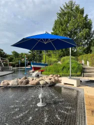 Ein schöner Außenbereich mit einem blauen Sonnenschirm und einem kleinen Brunnen. Im Hintergrund sind grüne Pflanzen und ein ruhiger Wasserbereich zu sehen.