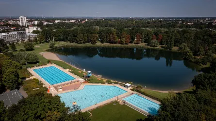 Ein Schwimmbad mit mehreren Becken liegt neben einem ruhigen See. Die Umgebung ist grün und von Bäumen umgeben.