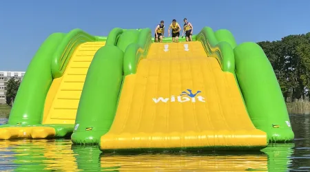 Eine große, grüne und gelbe Wasserrutsche steht an einem See. Drei Personen stehen oben und bereit zum Rutschen.