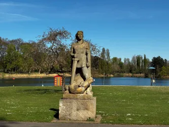 Eine Statue eines kriegerischen Mannes steht auf einem Podest. Im Hintergrund ist ein ruhiger See und Bäume zu sehen.