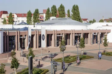 Ein modernes Einkaufszentrum mit einer großzügigen Außenterrasse. Umgeben von Bäumen und einem klaren Himmel.