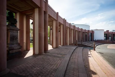 Ein architektonischer Innenhof mit Säulen und einem geschwungenen Weg. Im Hintergrund ist ein modernes Gebäude sichtbar, umgeben von klaren Himmel und Tageslicht.