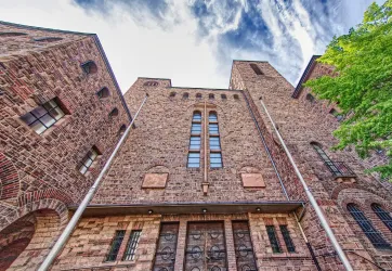 Ein historisches Gebäude aus Stein mit hohen Türmen und großen Fenstern. Die Szenerie wird von einem teilweise bewölkten Himmel und dem Grün von Bäumen ergänzt.
