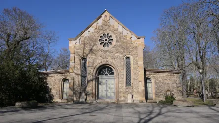 Ein historisches Gebäude aus Stein mit einem großen Rundfenster. Die Umgebung ist grün und die Bäume stehen ohne Blätter.