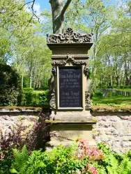Ein historischer Gedenkstein umgeben von Grün. Die Inschrift erinnert an verstorbene Personen.