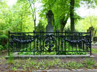 Ein kunstvoller eiserner Zaun umgibt ein Grab in einem ruhigen, grünen Friedhof. Überall sind hohe Bäume und das Licht filtert sanft durch die Blätter.