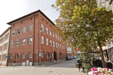 Ein rotes Gebäude an einer Straßenecke, umgeben von Bäumen und Pflanzen. Die Straße wirkt ruhig und urban.