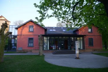 Ein rotes Gebäude mit modernen Elementen und großen Fenstern. Der Eingang ist von einer Wiese und Bäumen umgeben.
