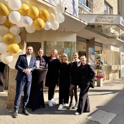 Eine Gruppe von sechs Personen steht vor einem Schmuckgeschäft mit einer festlichen Dekoration aus Luftballons. Das Schaufenster zeigt verschiedene Schmuckstücke und die Sonne scheint.