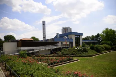 Ein modernes Gebäude mit einem blauen Dach und vielen Fenstern. Im Vordergrund befindet sich ein schön angelegter Garten mit Blumen und Bäumen.