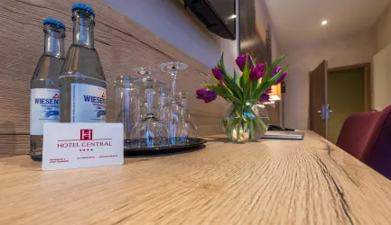 Ein eleganter Tisch in einem Hotelzimmer mit zwei Flaschen Wasser, Gläsern und einem Blumengesteck. Im Vordergrund liegt eine Zimmerschlüsselkarte von Hotel Central.