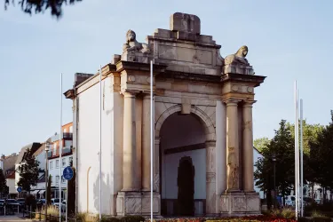 Ein historisches Tor mit Statuen an den Seiten. Die Umgebung ist grün und belebt.