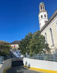 Ein Zugang zu einer Tiefgarage mit einem Sichtfeld auf einen Kirchturm. Im Hintergrund sind Bäume und ein klarer blauer Himmel zu sehen.