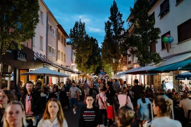 Eine belebte Einkaufsstraße bei Dämmerung mit vielen Menschen. Kioske und Restaurants laden zum Verweilen ein.