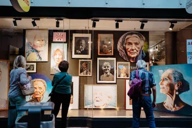 Eine Kunstgalerie mit verschiedenen Porträts an der Wand. Drei Personen betrachten die Gemälde aufmerksam.