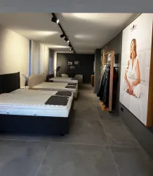 Ein modernes Schlafzimmergeschäft mit mehreren Betten auf einer Fliese. An der Wand hängt ein großes Werbeschild mit einem Modell, das eine Matratze präsentiert.