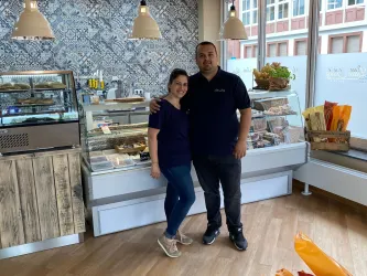 Ein Paar steht in einem gemütlichen Café mit einem süßen Angebot im Hintergrund. Beide tragen dunkelblaue T-Shirts und lächeln freundlich.