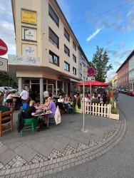 Ein gemütliches Bistro mit Außenterrasse, in dem viele Menschen speisen. Umgeben von einer freundlichen Straße und Gebäuden in Stadtatmosphäre.