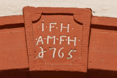 Ein historisches Wappen mit der Inschrift "IFH AM FH 1765" ist an einer roten Wand angebracht. Das Design ist schlicht und zeigt handwerkliche Details.