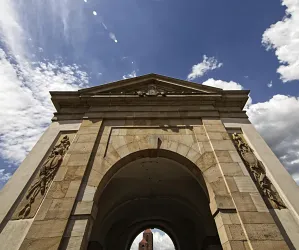 Ein großer Torbogen aus Stein unter einem blauen Himmel mit weißen Wolken. Die Fassade ist mit kunstvollen Verzierungen geschmückt.