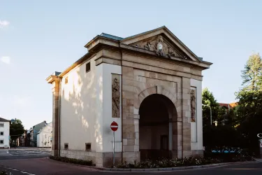 Ein historisches Tor mit einem großen Rundbogen und dekorativen Elementen. Es befindet sich an einer Straßenkreuzung und ist von Bäumen umgeben.