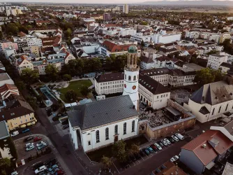 Eine Luftaufnahme einer Stadt mit vielen Gebäuden und einem Uhrenturm. Im Vordergrund sind Autos und ein parkähnlicher Bereich zu sehen.