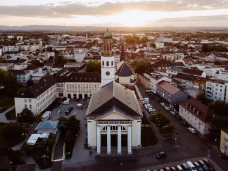 Eine Stadtansicht mit einer Kirche im Vordergrund und einer Wohngegend im Hintergrund. Die Sonne geht am Horizont unter und schafft eine warme Atmosphäre.