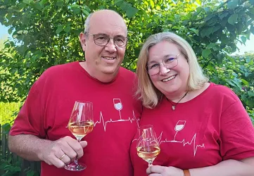 Ein Paar hält Weingläser und lächelt glücklich. Sie tragen rote T-Shirts mit einem lustigen Design und stehen in einem grünen Garten.