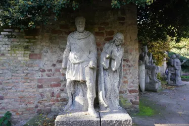 Zwei große Steinskulpturen stehen an einer Mauer. Die Figuren zeigen einen Mann und eine Frau in antiker Kleidung.