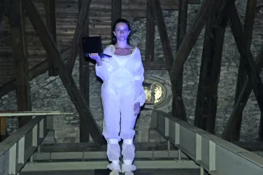 Eine Person in einem weißen Schutzanzug steht in einem Raum mit sichtbaren Holzstrukturen. Sie hält ein schwarzes Objekt in der Hand und ist mit blauem Licht beleuchtet.