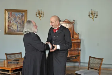 Zwei Männer in historischen Anzügen schütteln sich die Hände in einem eleganten Raum. Im Hintergrund sieht man ein Gemälde und ein Möbelstück aus Holz.
