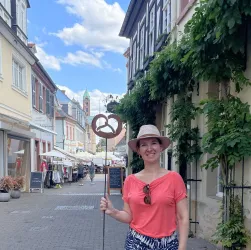 Eine Frau hält ein Schild mit einem Brezel-Motiv in einer schönen Straße. Im Hintergrund sind Geschäfte und blauem Himmel zu sehen.