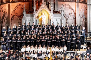 Ein großer Chor steht in einer Kirche und singt. Im Hintergrund ist eine beeindruckende Statue und farbenfrohes Licht zu sehen.